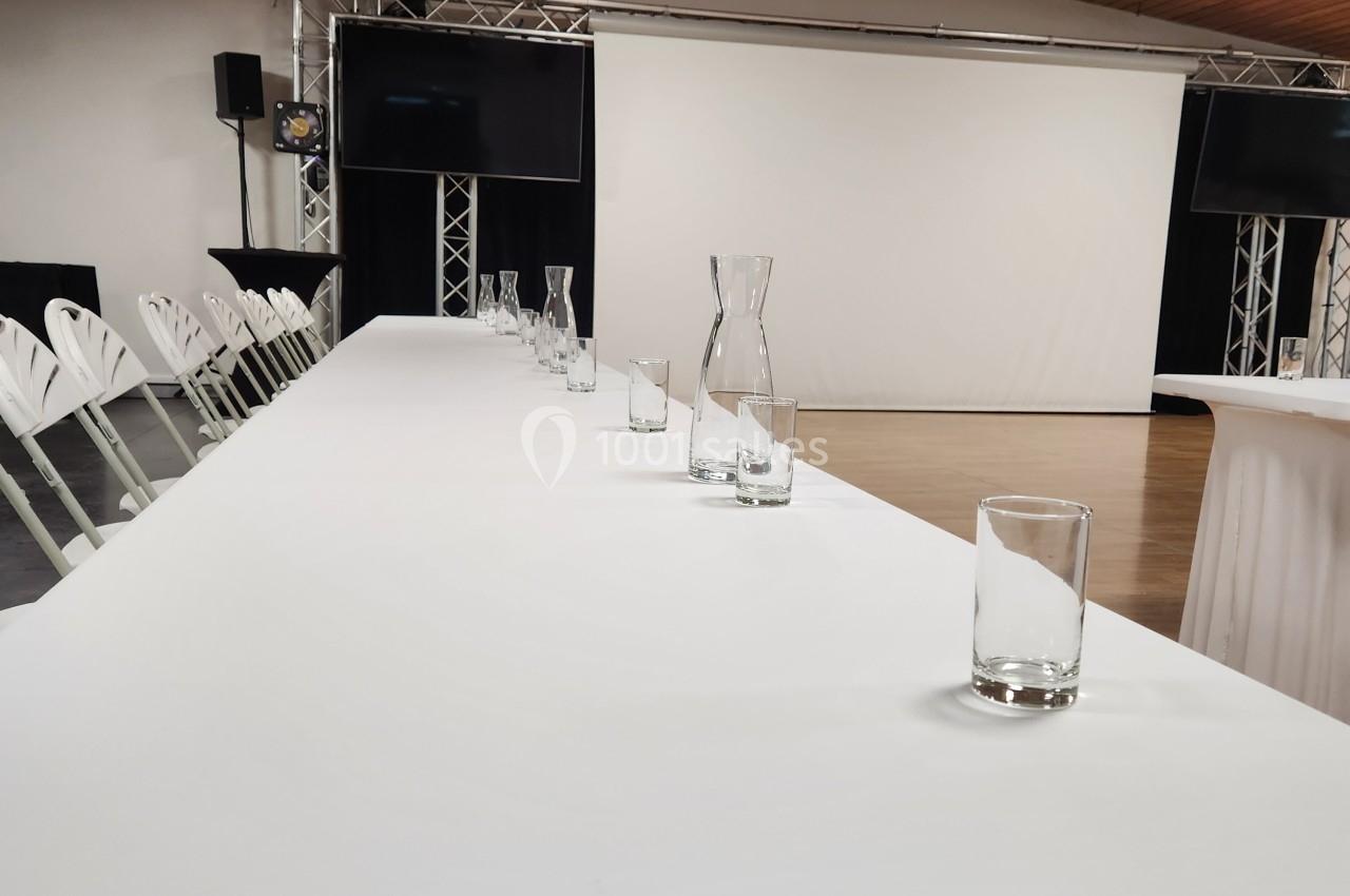 Salle de conférence avec une grande table blanche, des verres et des carafes, face à un écran de projection.