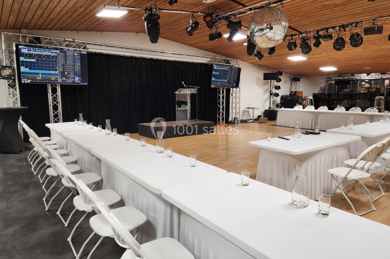 Salle de conférence aménagée avec des tables blanches, des chaises, un pupitre et des écrans pour une présentation.