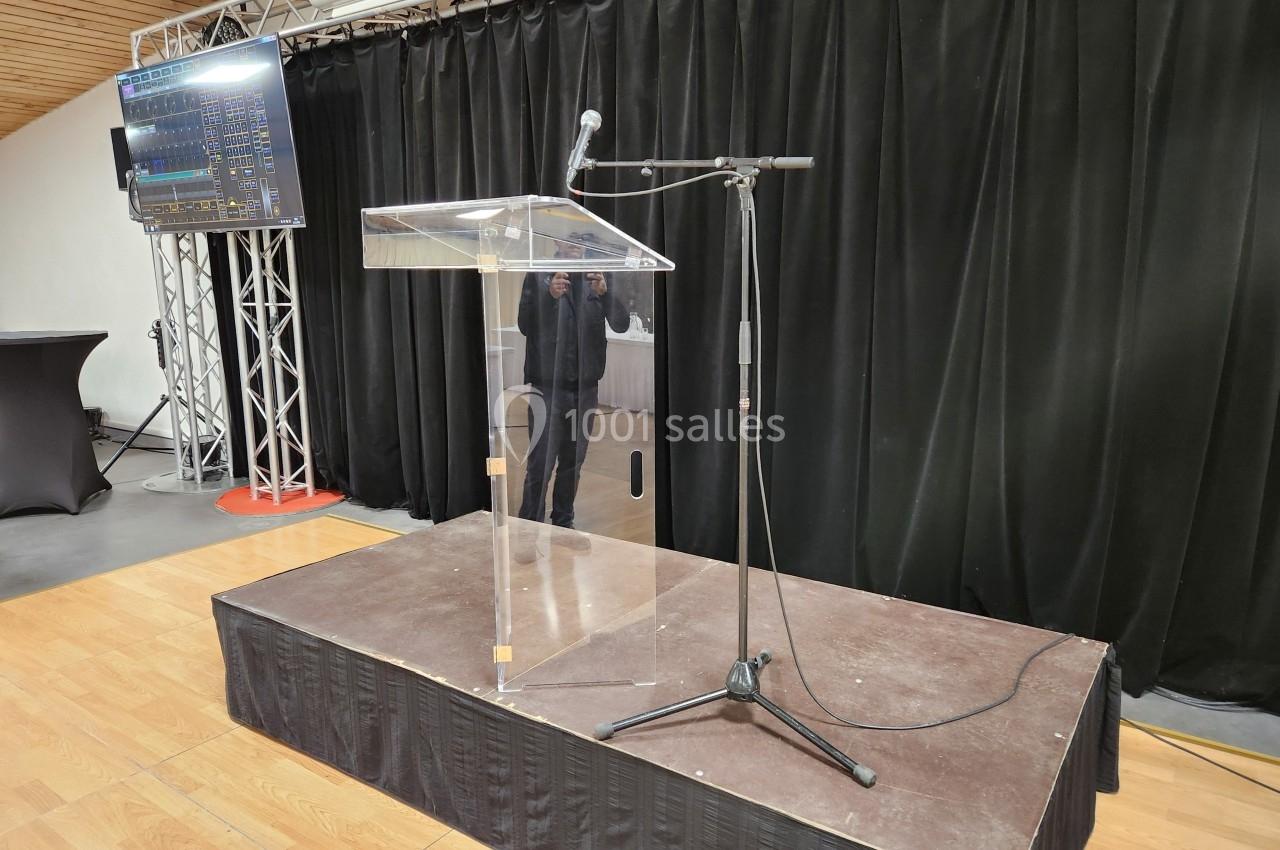 Un pupitre transparent avec microphone sur une petite estrade en bois, devant un rideau noir et un écran latéral.