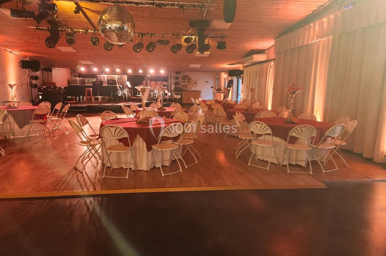 Salle de réception décorée avec des tables rondes nappées de rouge, éclairage chaleureux et scène en arrière-plan.