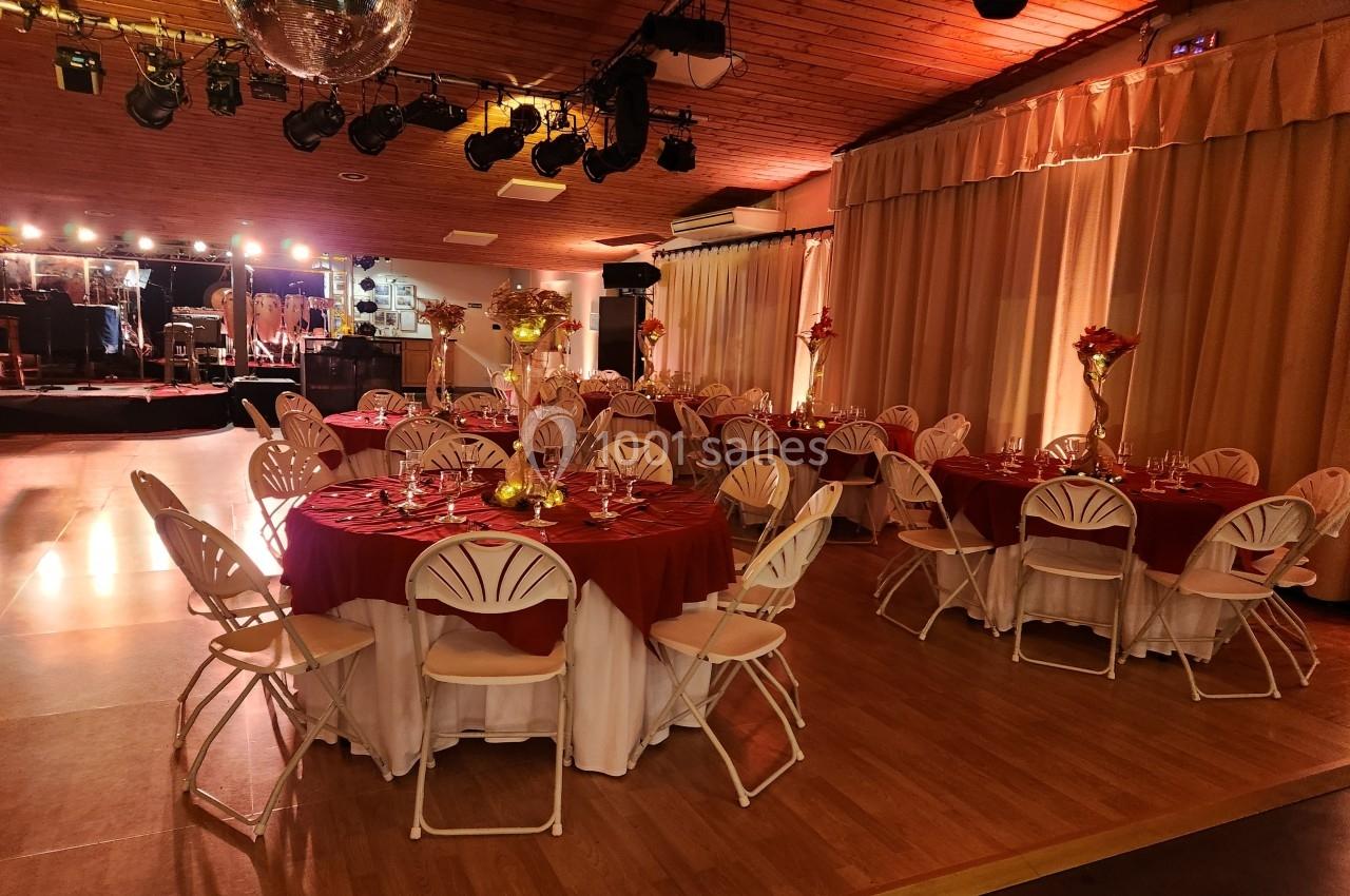 Salle de réception décorée avec des tables rondes, nappes rouges, chaises blanches et éclairage chaleureux.