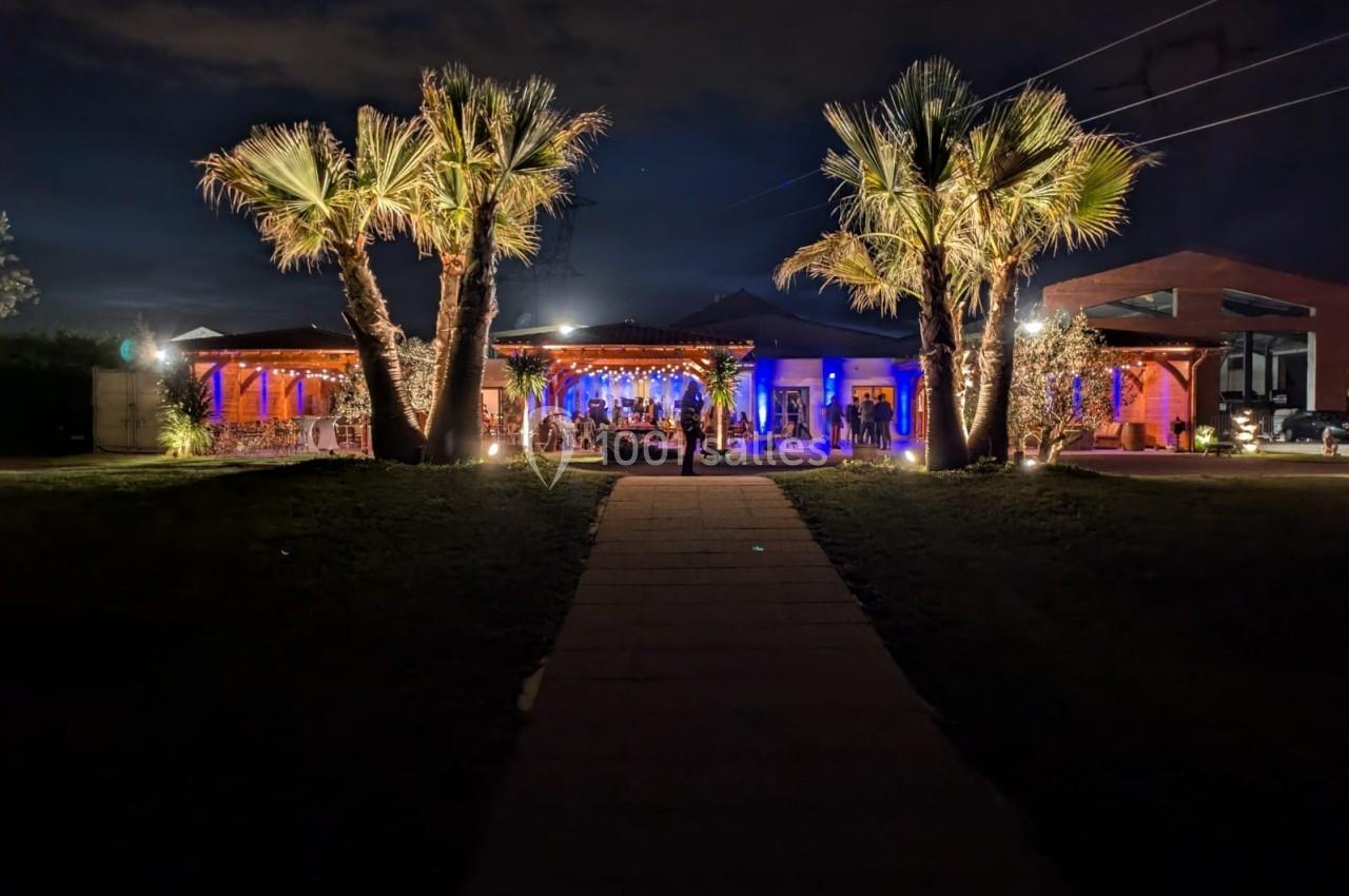 Allée menant à un bâtiment éclairé de lumières colorées, entouré de palmiers dans un cadre nocturne.