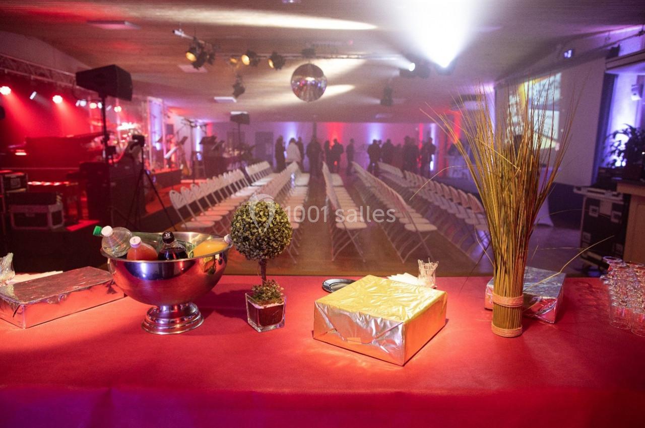 Salle de réception avec rangées de chaises, éclairage coloré, table au premier plan décorée et garnie d'objets divers.