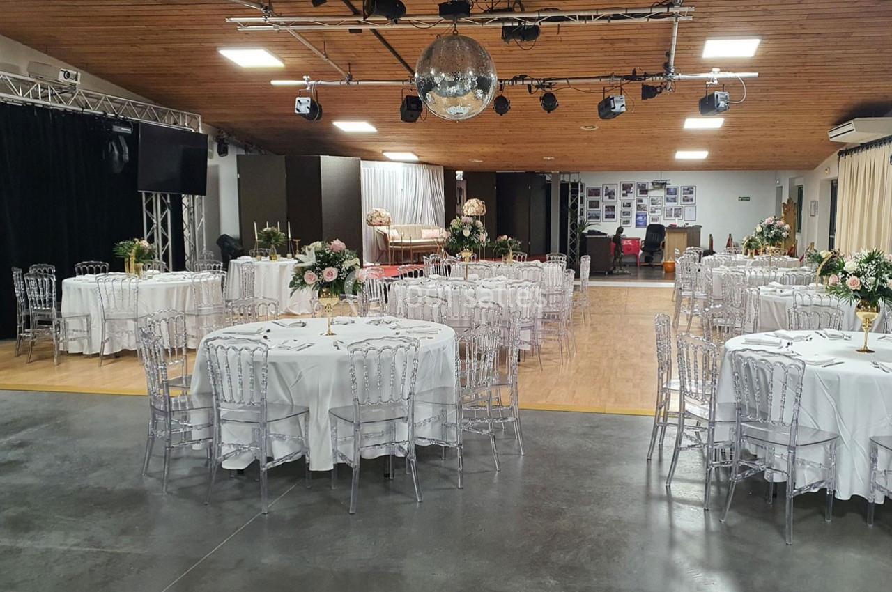 Salle de réception décorée avec des tables rondes, nappes blanches, chaises transparentes et arrangements floraux.