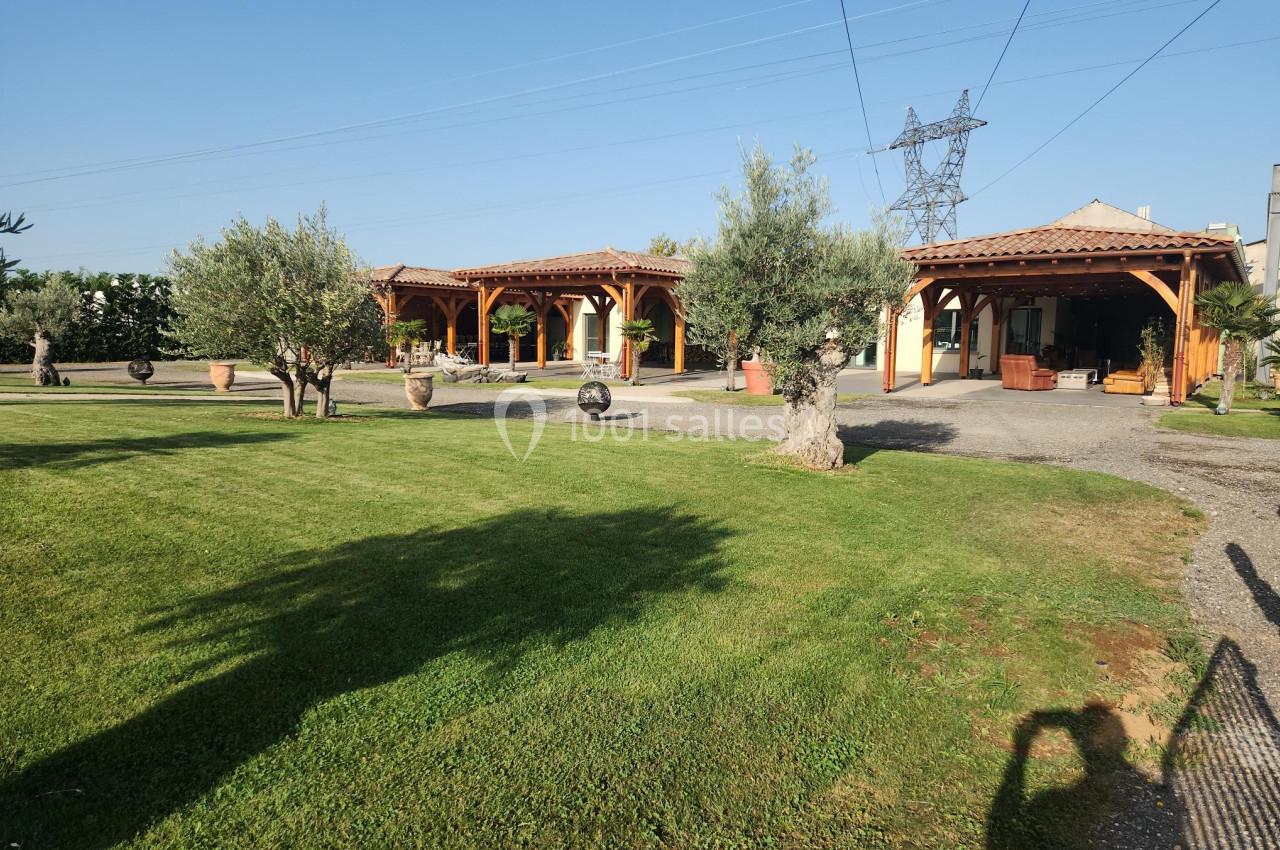 Vue d'un jardin avec pelouse, oliviers, et bâtiments à toit en tuiles sous un ciel dégagé.