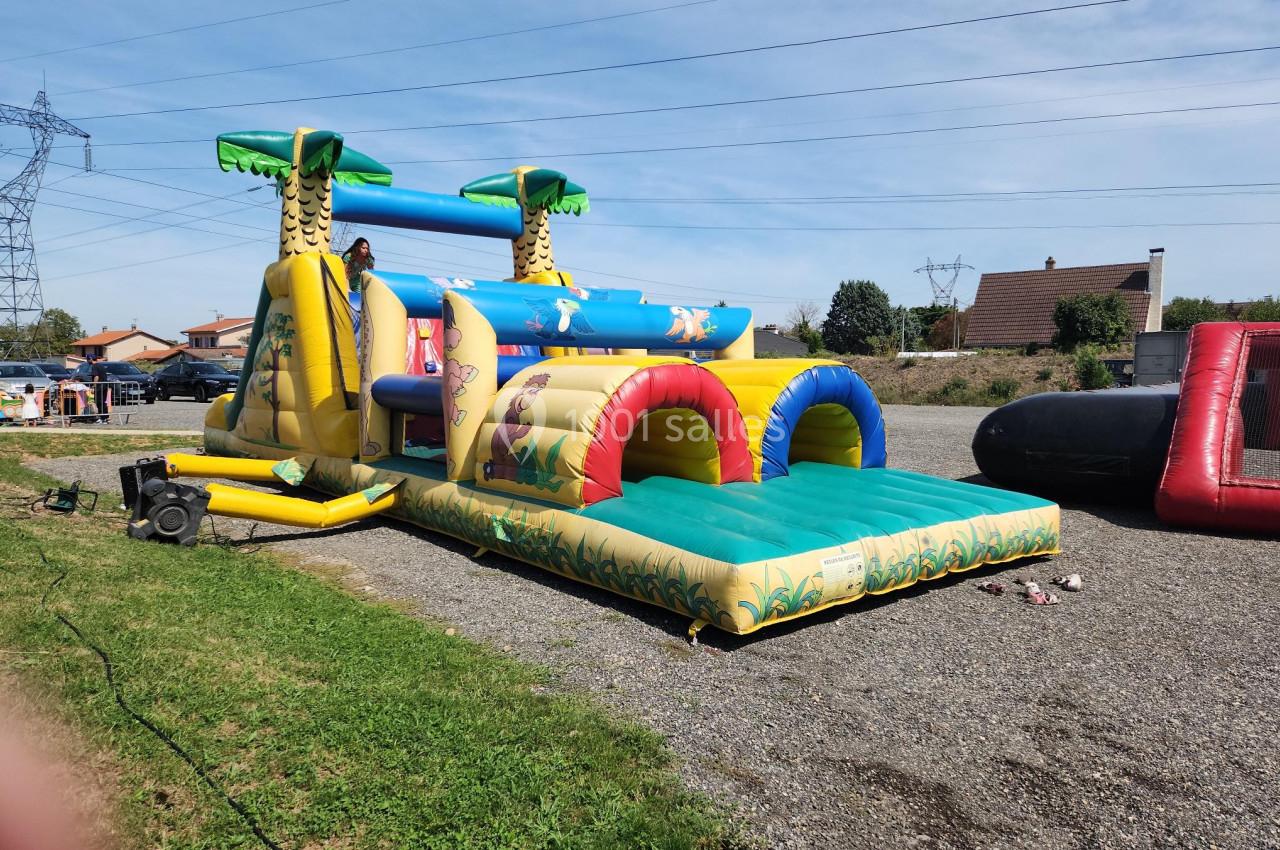 Structure gonflable colorée avec toboggan et tunnels, installée en extérieur sur un sol gravillonné près d'une pelouse.
