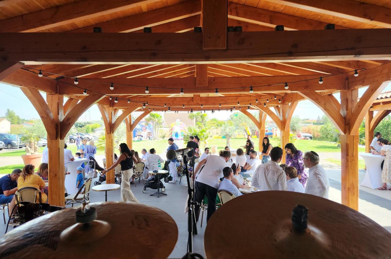 Vue depuis une batterie sous une pergola en bois, avec des invités assis et debout lors d'un événement en plein air.