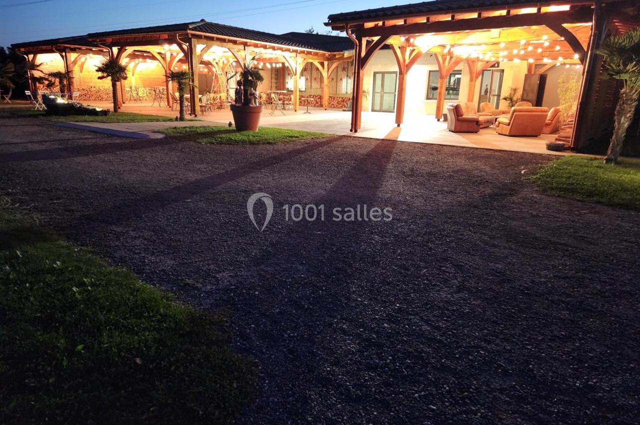 Vue nocturne d'une terrasse couverte éclairée, entourée de verdure et d'une allée en gravier.
