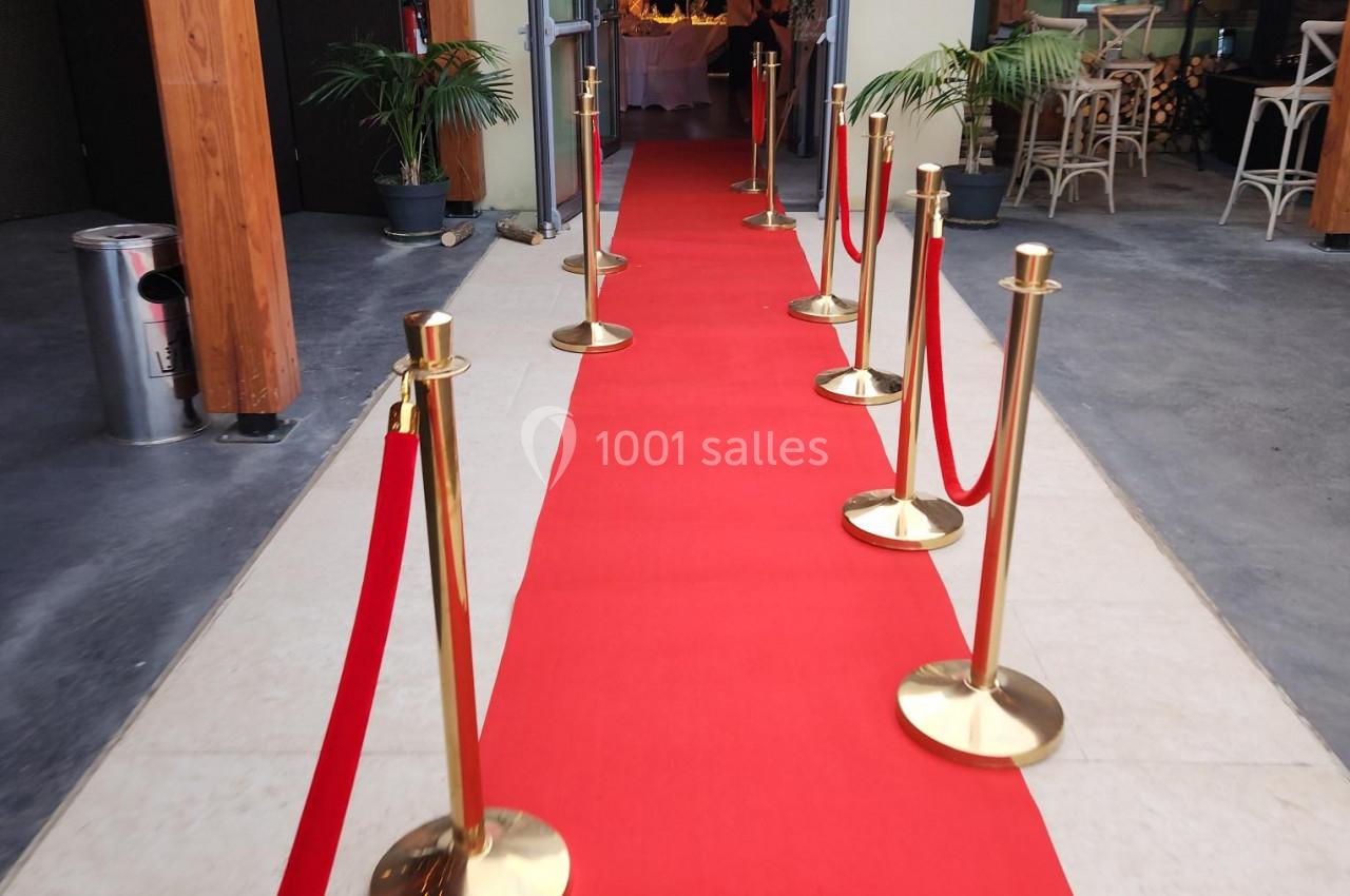 Tapis rouge bordé de poteaux dorés menant à une entrée ouverte dans un bâtiment moderne avec des plantes décoratives.