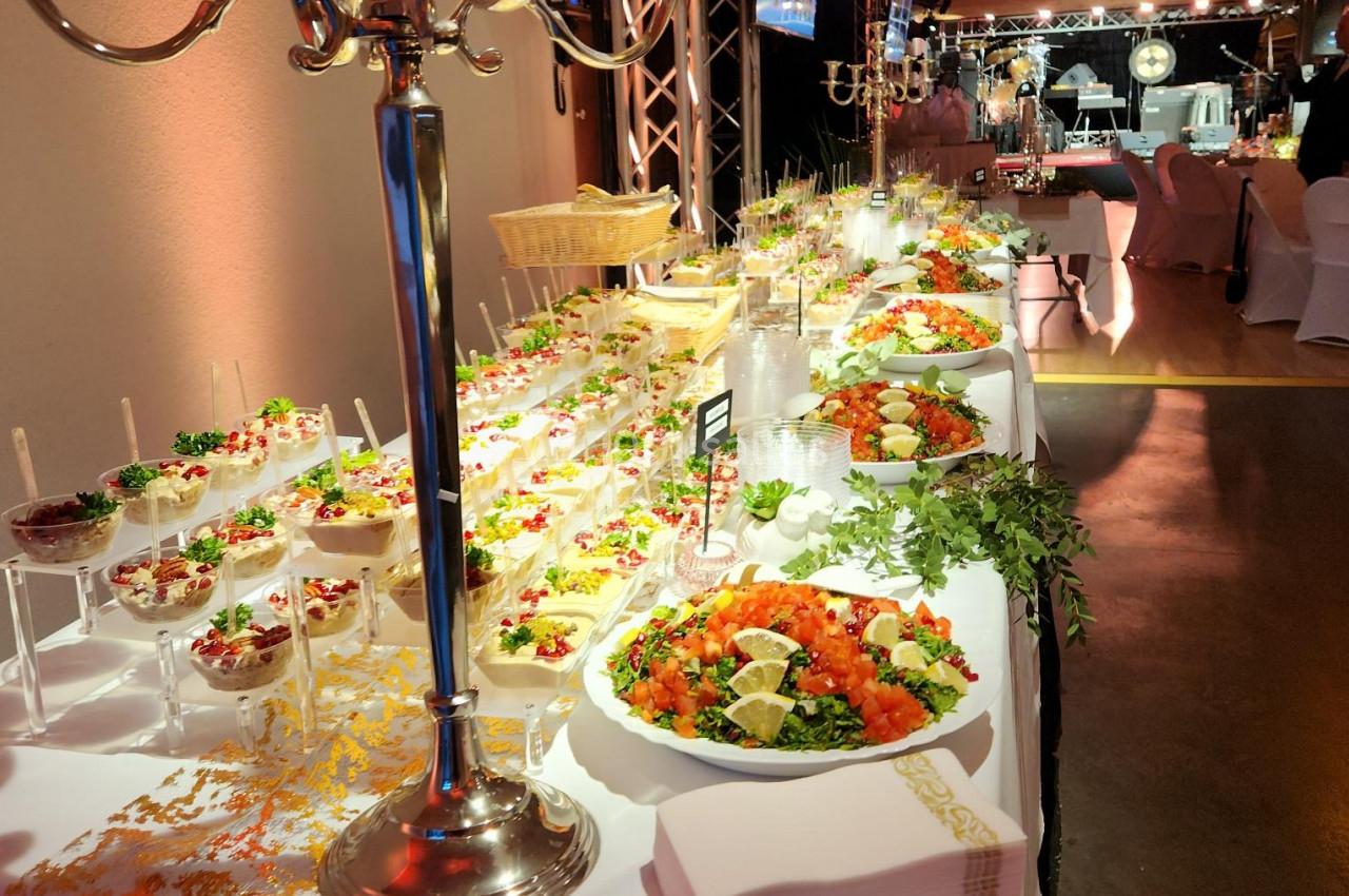 Buffet varié avec plats colorés, verrines, salades et décorations élégantes dans une salle de réception éclairée.