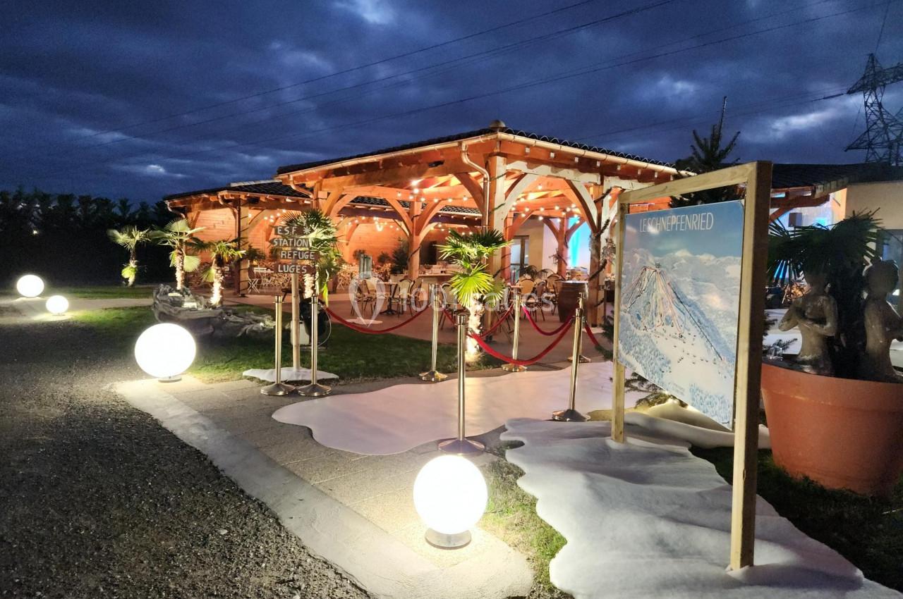 Chalet en bois éclairé la nuit, entouré de décorations lumineuses et d'une affiche montrant un paysage enneigé.