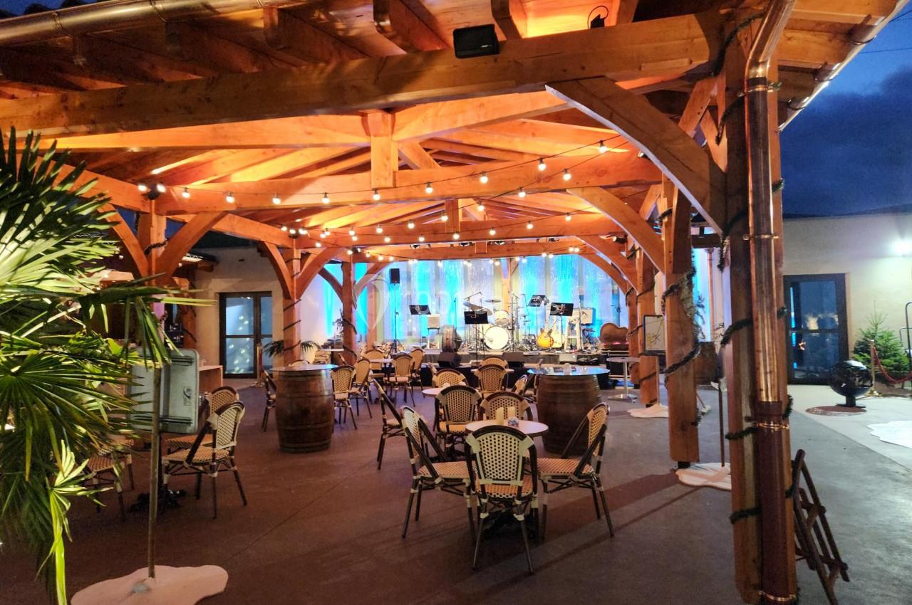 Structure en bois éclairée avec des guirlandes lumineuses, tables et chaises disposées pour un événement en soirée.