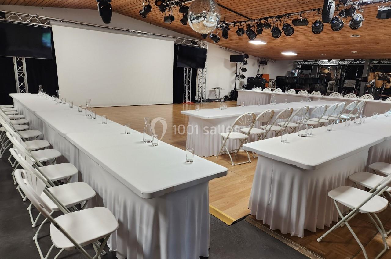 Salle de réunion aménagée avec des tables blanches en U, des chaises pliantes et un écran de projection en fond.