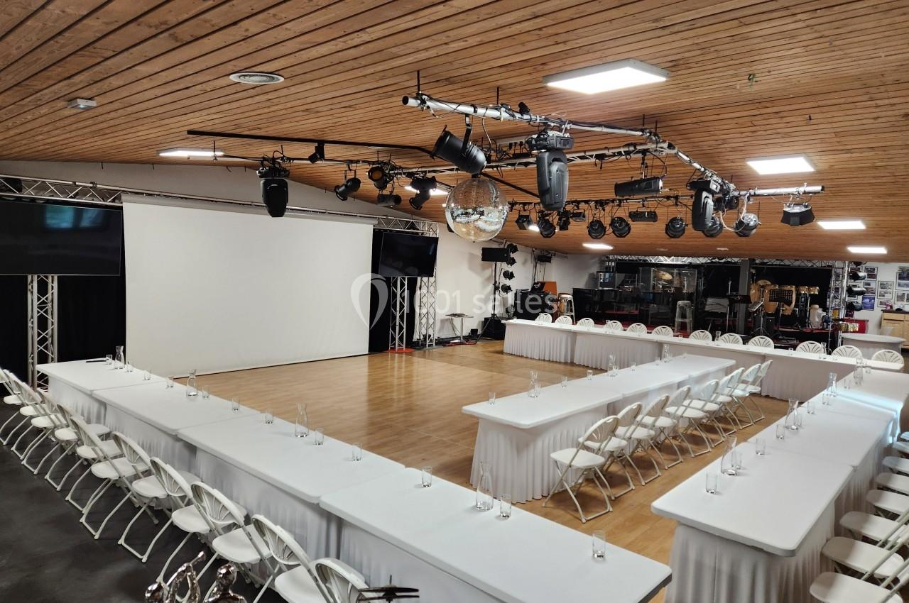 Salle de conférence avec tables en U, chaises blanches, écran de projection, éclairages suspendus et parquet au sol.