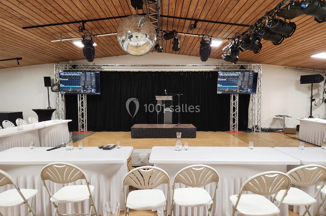 Salle de conférence avec estrade, pupitre, écrans, tables blanches et chaises alignées sous un plafond en bois.
