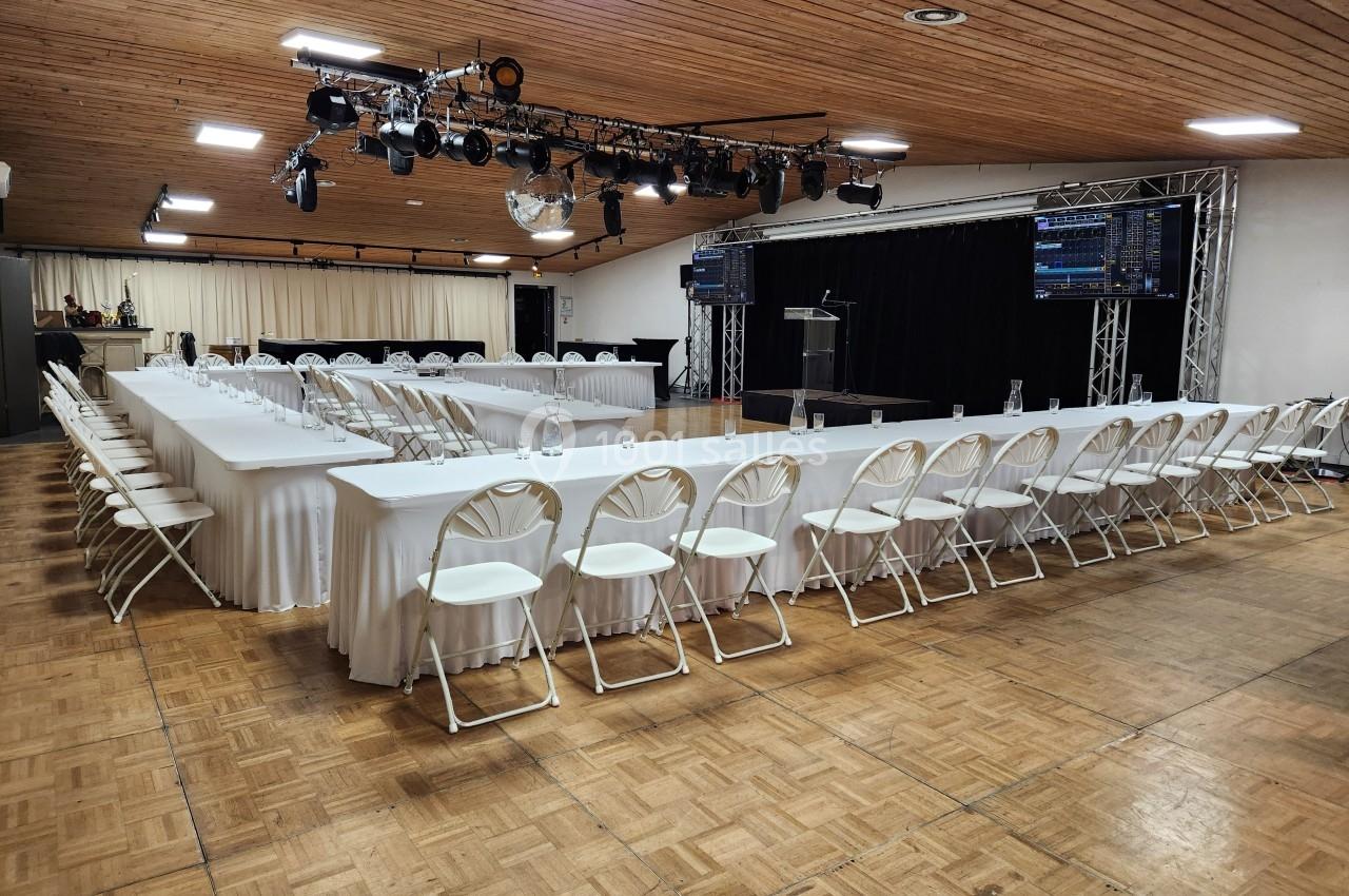 Salle de conférence avec des tables en U, des chaises blanches, un podium et des équipements audiovisuels.