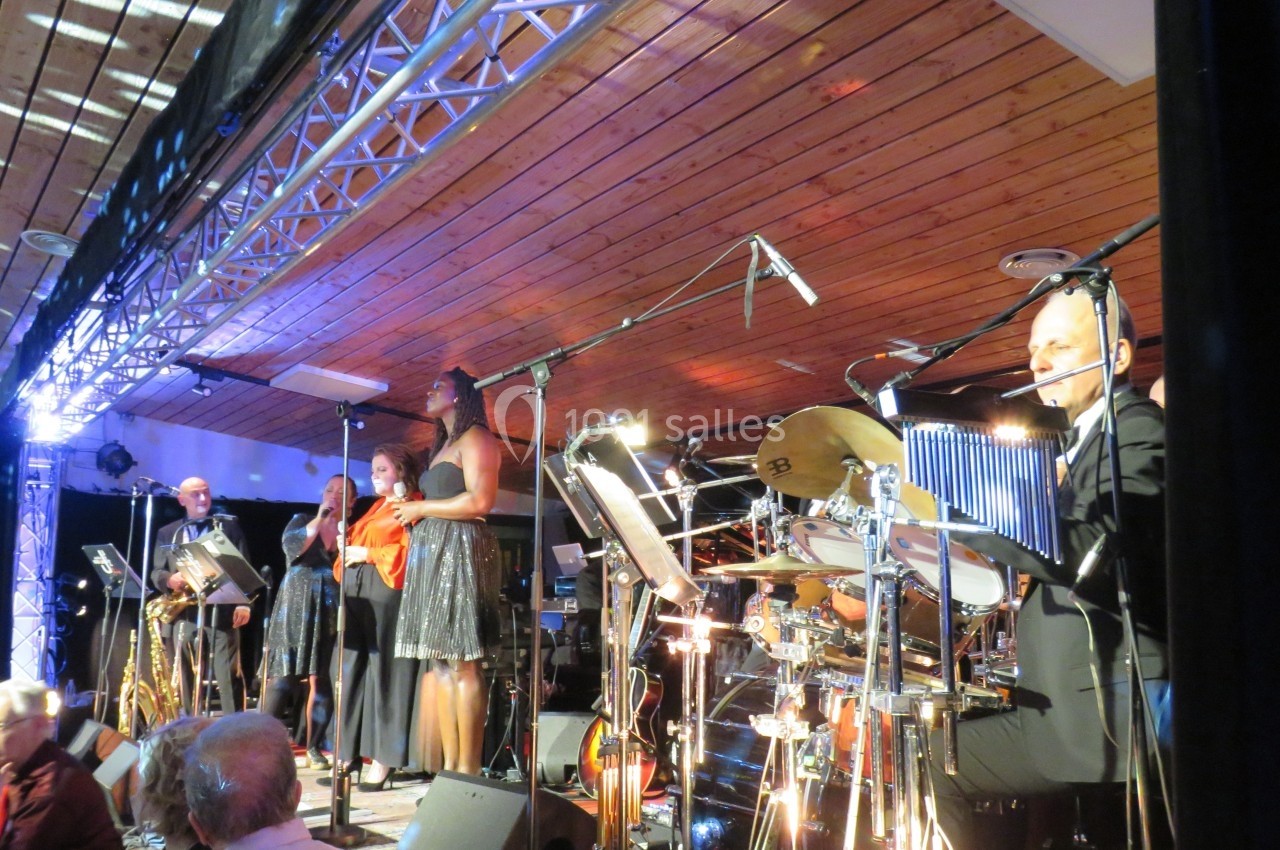 Un groupe de musiciens et chanteurs se produit sur une scène éclairée sous un plafond en bois.