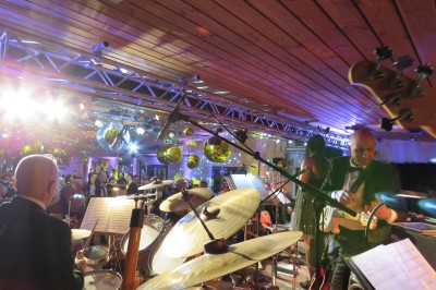 Un groupe de musiciens et chanteurs joue sur une scène éclairée dans une salle avec un public assis.