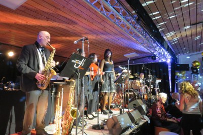 Un groupe de musiciens et chanteurs joue sur une scène éclairée dans une salle avec un public assis.