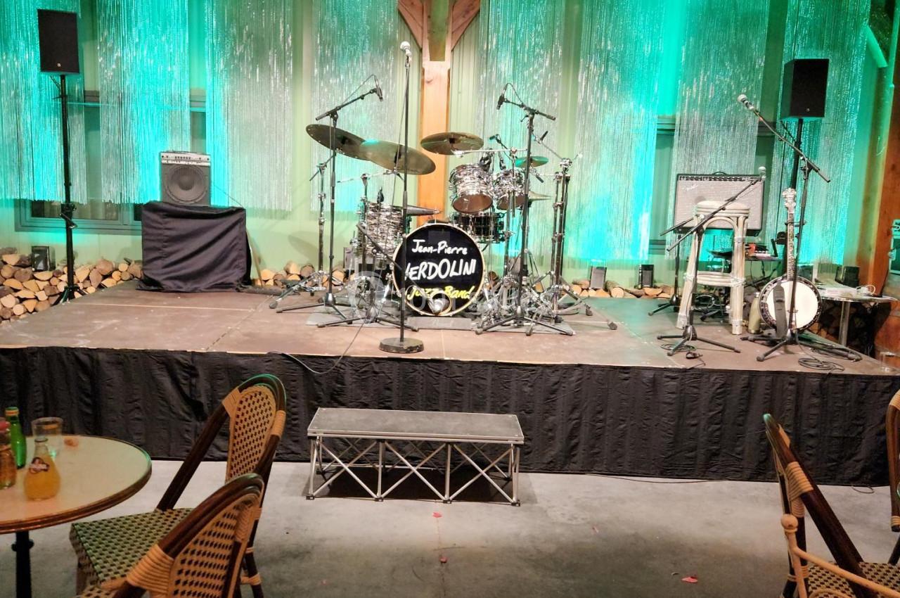 Scène vide avec une batterie, des amplificateurs et des éclairages verts, entourée de chaises et de tables en bois.