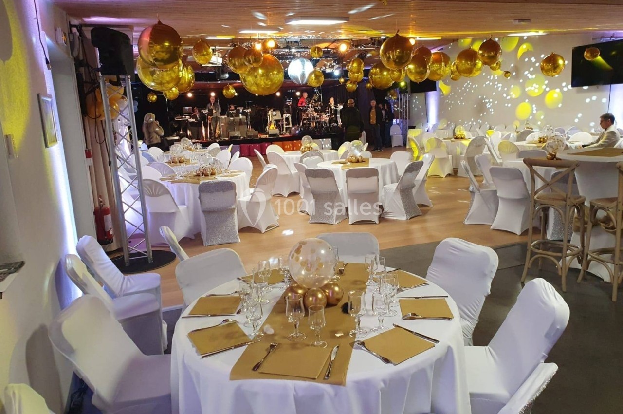 Salle décorée pour un événement avec tables rondes dressées, chaises blanches et ballons dorés suspendus.