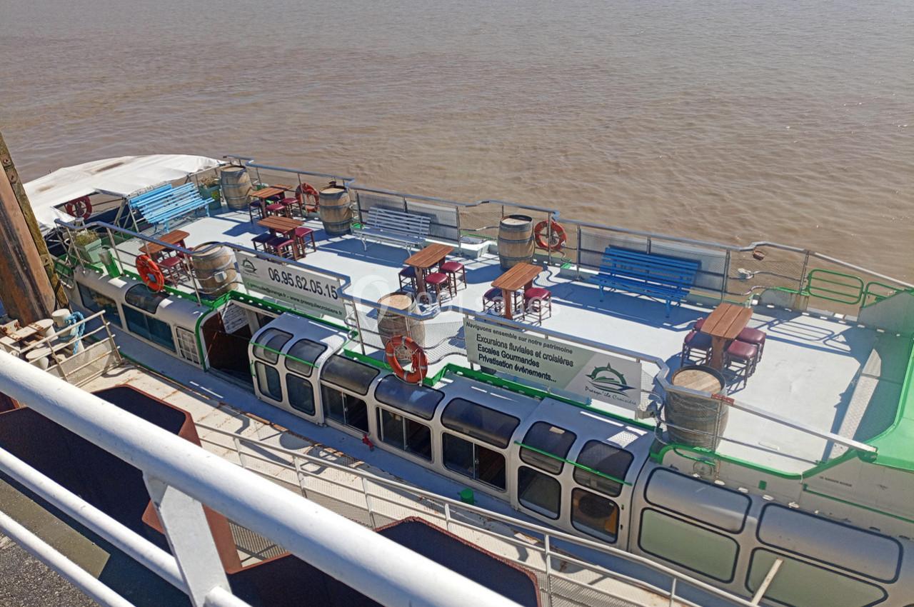 Bateau à pont ouvert avec bancs, tables et bouées de sauvetage, amarré au bord d'une rivière.