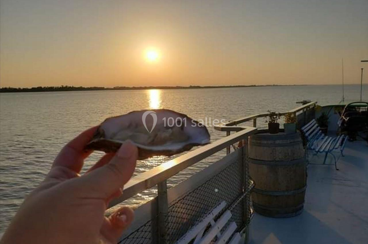 Une main tient une huître ouverte sur un bateau au coucher du soleil, avec vue sur l'eau calme et l'horizon.