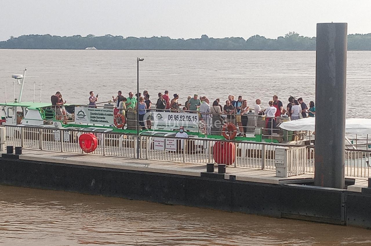 Des passagers attendent sur un bateau amarré à un ponton au bord d'un large fleuve entouré de végétation.