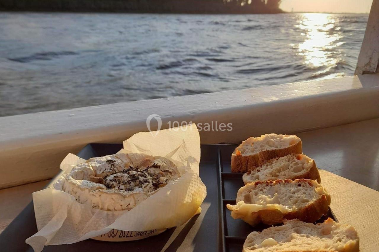 Fromage coulant et tranches de pain sur un plateau noir, posés près d'une fenêtre avec vue sur un lac au coucher du soleil.