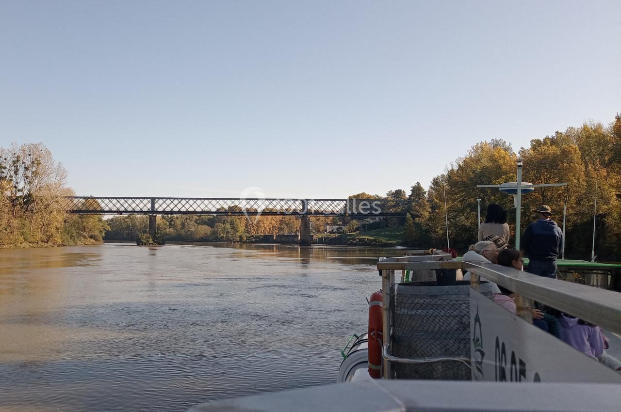 Vue depuis un bateau sur une rivière, avec un pont métallique à l'horizon et des arbres bordant les rives.