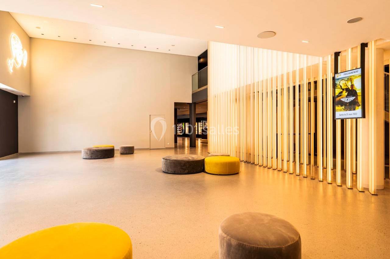 Hall spacieux avec sol clair, poufs jaunes et gris, murs minimalistes, et panneaux lumineux verticaux.