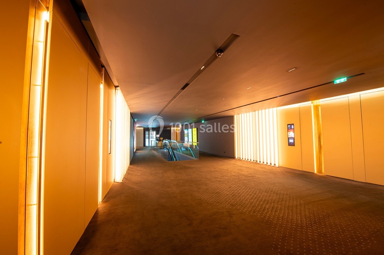 Couloir moderne avec éclairage chaleureux, murs clairs et tapis sombre, menant à une zone vitrée illuminée.
