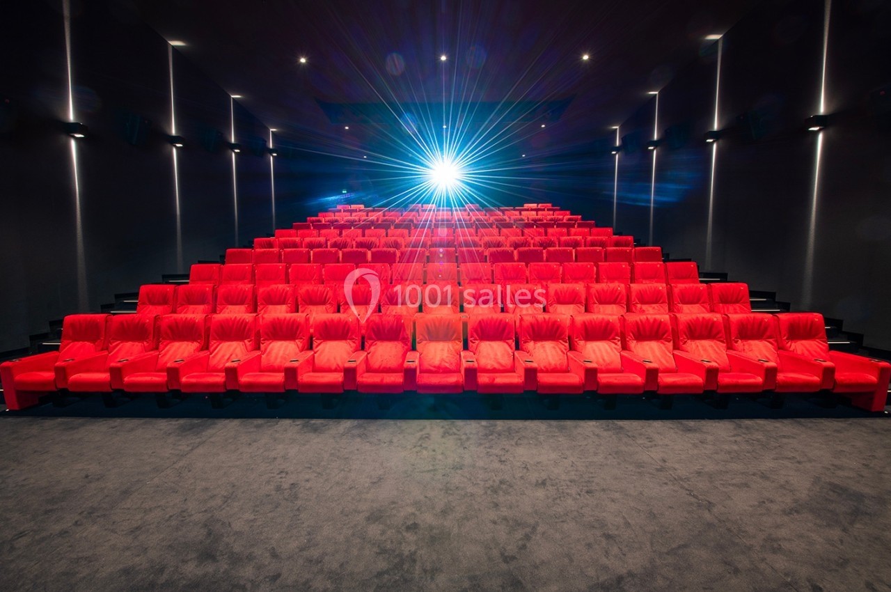 Salle de cinéma vide avec des rangées de fauteuils rouges et un projecteur allumé au fond.