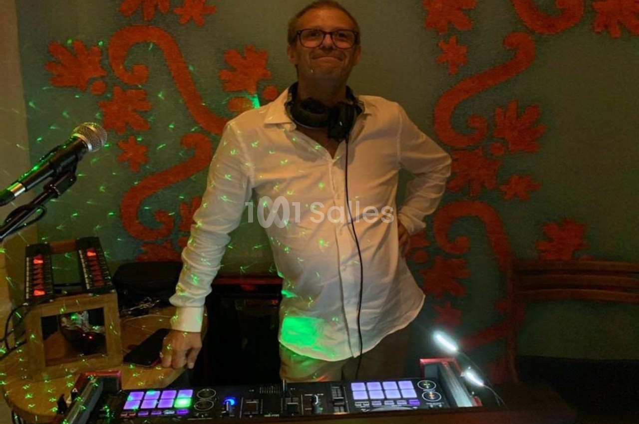 Homme debout derrière une table de mixage éclairée, portant un casque audio, avec un fond décoratif et des lumières colorées.