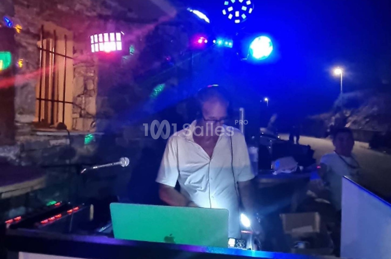 Un DJ mixe en extérieur de nuit, entouré de lumières colorées et d'équipements audio.