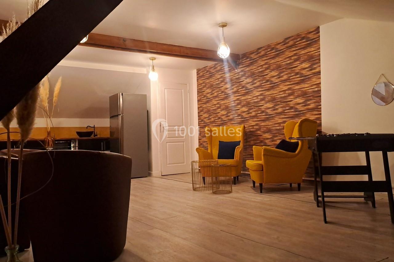 Espace intérieur avec fauteuils jaunes, table basse, baby-foot, réfrigérateur et décoration murale en bois.