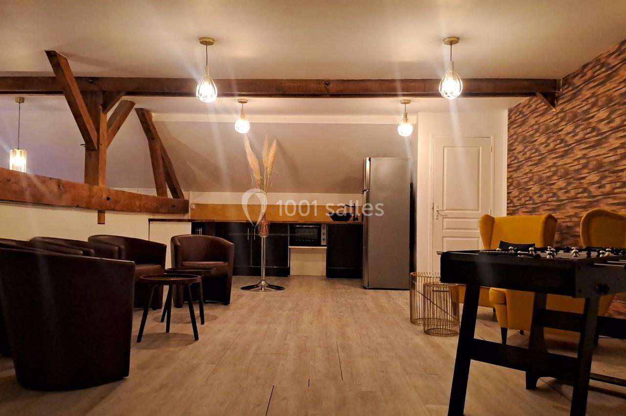 Espace aménagé avec coin salon, cuisine équipée, table de baby-foot et éclairage suspendu dans une pièce mansardée.