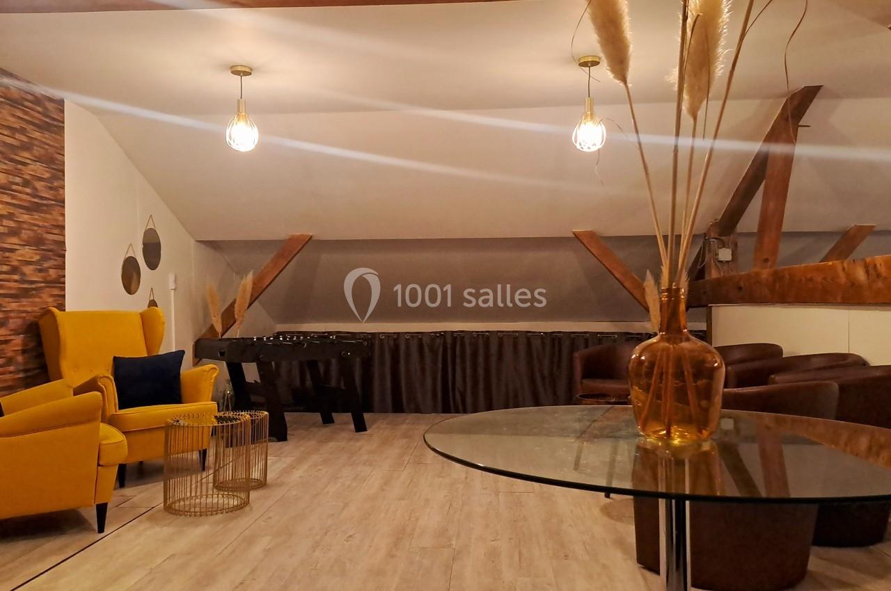 Espace salon mansardé avec fauteuils, table en verre, baby-foot et décoration moderne sous poutres apparentes.