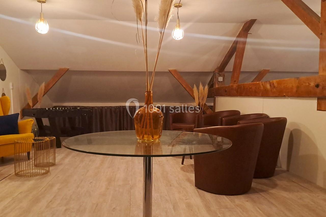 Salle mansardée avec table en verre, fauteuils en cuir marron, fauteuil jaune et décoration minimaliste.