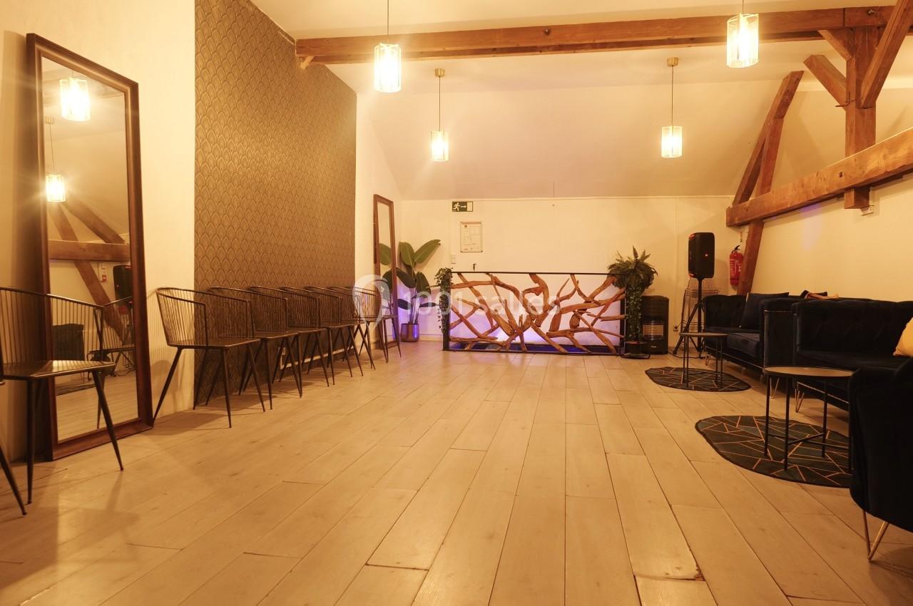 Salle lumineuse avec parquet clair, chaises alignées, canapé, plantes et comptoir en bois sculpté.