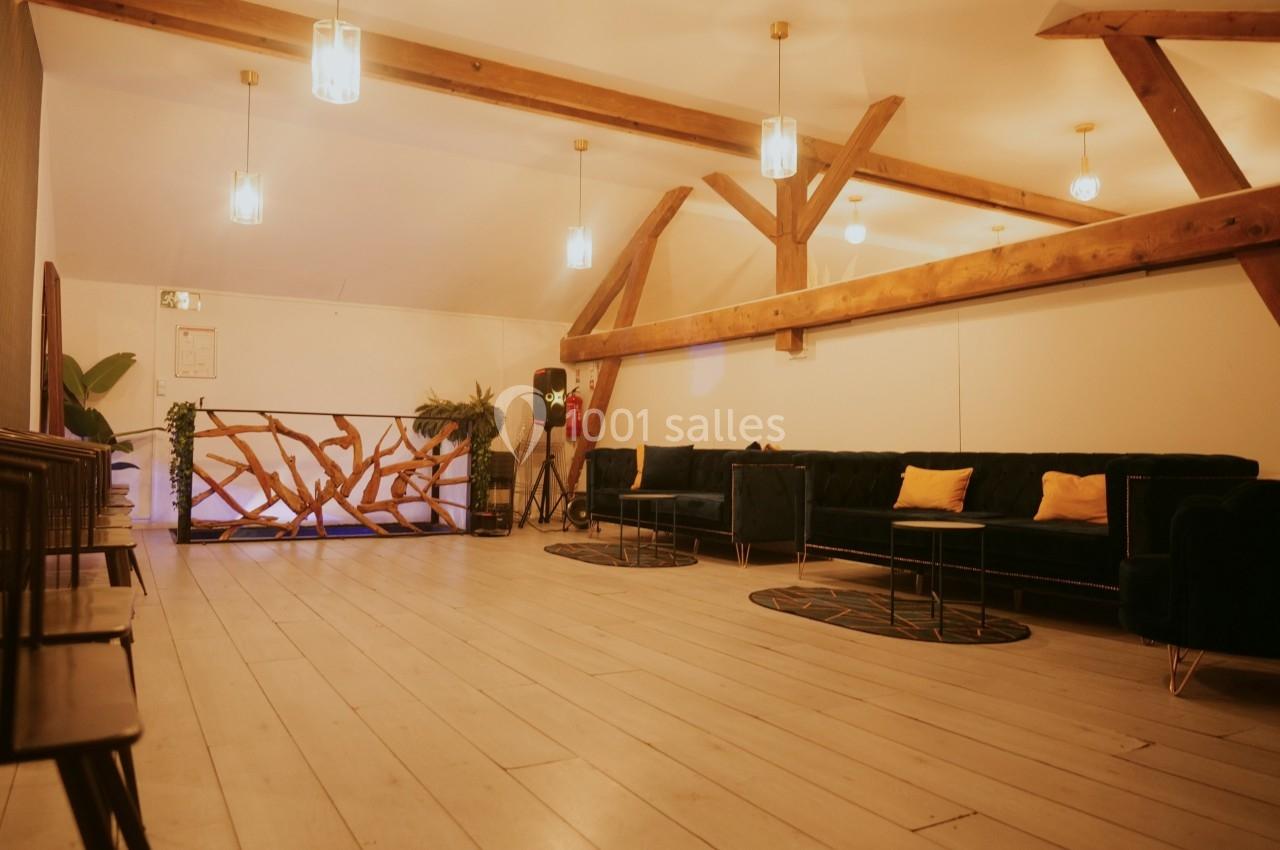 Salle lumineuse avec poutres apparentes, canapés bleus, coussins jaunes, plantes et sol en bois clair.