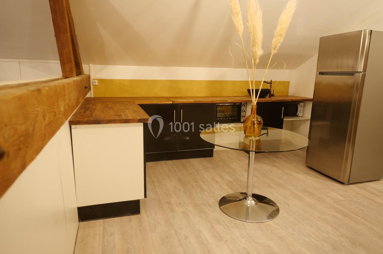 Cuisine sous combles avec meubles noirs, plan de travail en bois, table ronde en verre et réfrigérateur en inox.