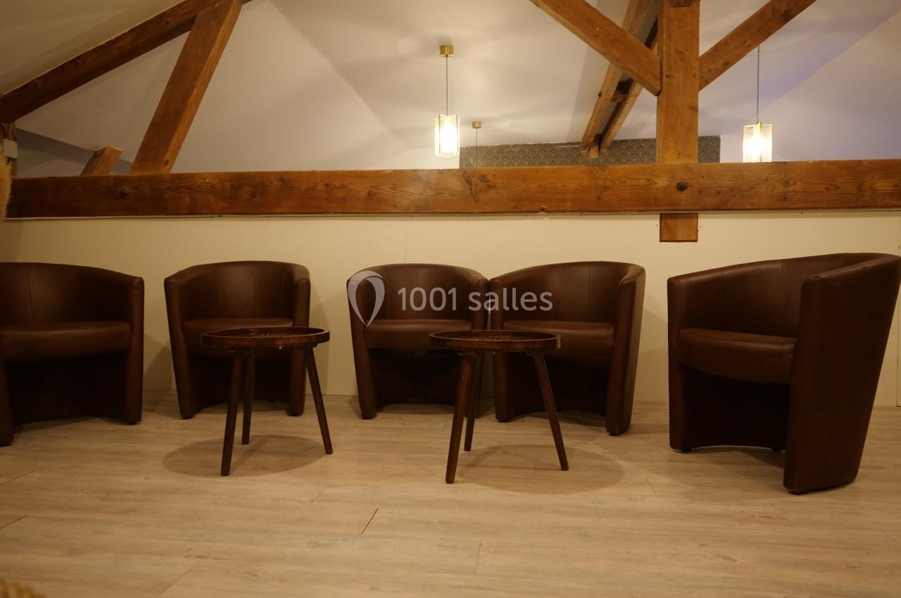 Quatre fauteuils en cuir marron disposés autour de deux petites tables en bois dans une pièce avec poutres apparentes.