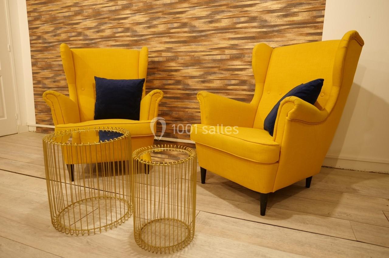 Deux fauteuils jaunes avec coussins bleus, accompagnés de deux tables basses dorées, sur un sol en bois clair.