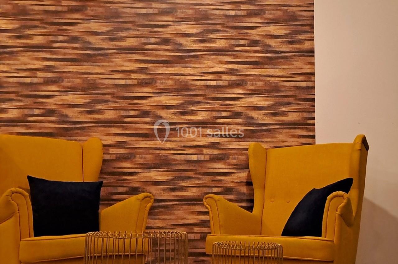 Deux fauteuils jaunes avec coussins noirs entourent deux tables basses dorées, devant un mur texturé en bois.