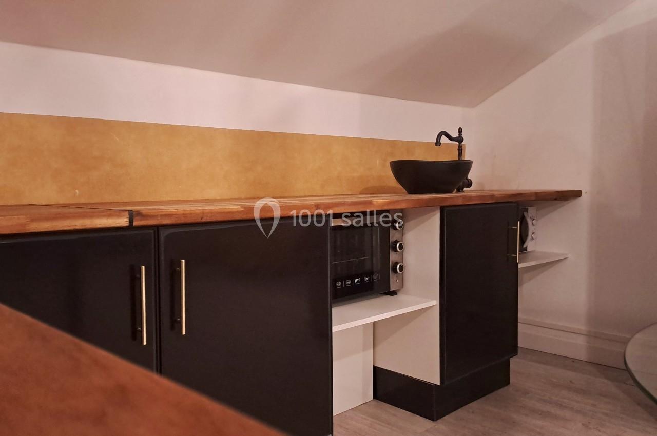 Cuisine sous pente avec plan de travail en bois, évier noir, placards noirs et crédence jaune.