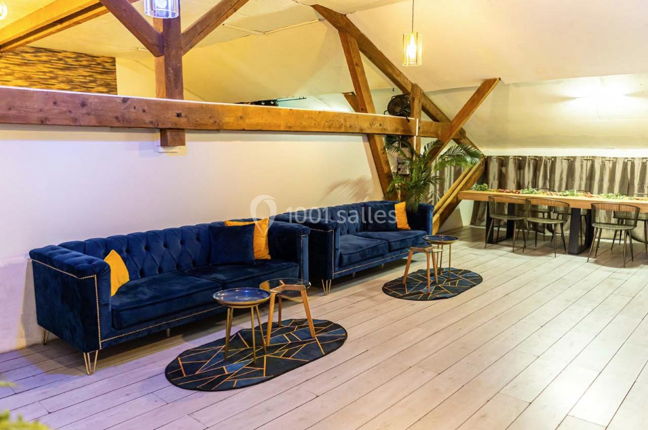 Deux canapés bleus avec coussins jaunes, tables basses et tapis géométriques dans un espace lumineux avec poutres apparentes.