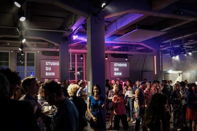 Un DJ mixe sur une scène éclairée par des lumières colorées, face à une foule dansante dans une salle sombre.
