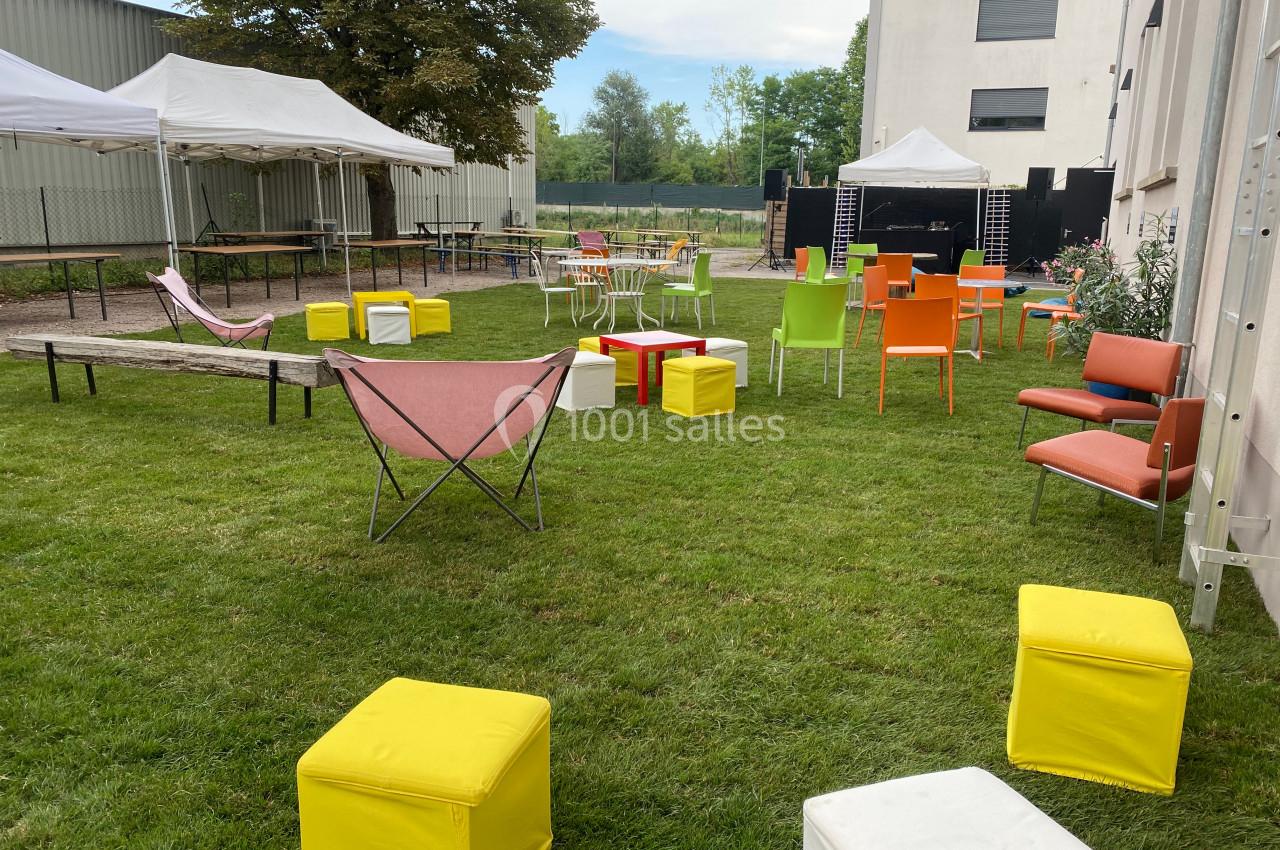 Espace extérieur avec pelouse aménagé de chaises colorées, tables, poufs et tentes blanches en arrière-plan.