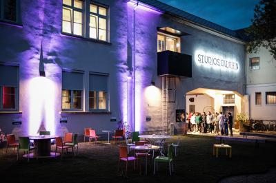 Un DJ mixe sur une scène éclairée par des lumières colorées, face à une foule dansante dans une salle sombre.